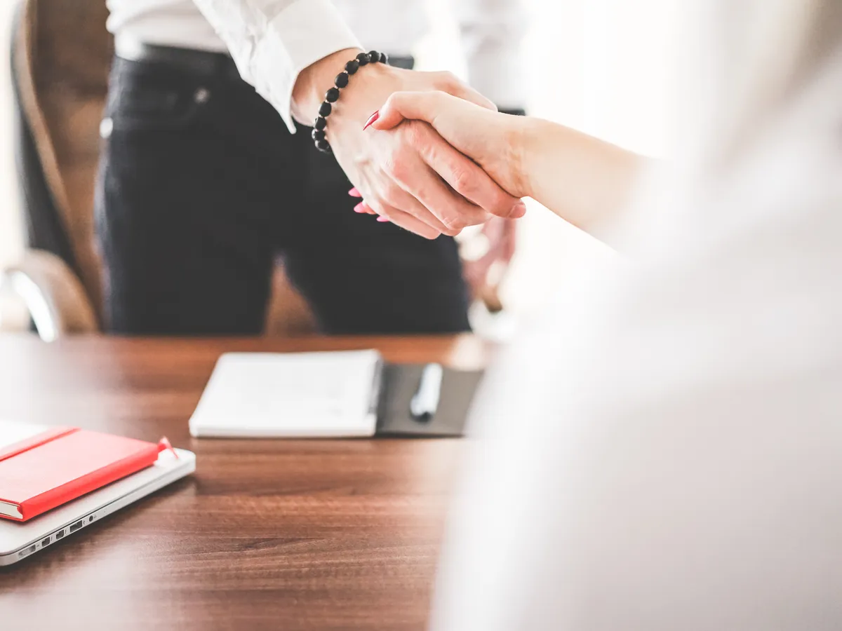 Two people shaking hands across a desk with a notebook and laptop