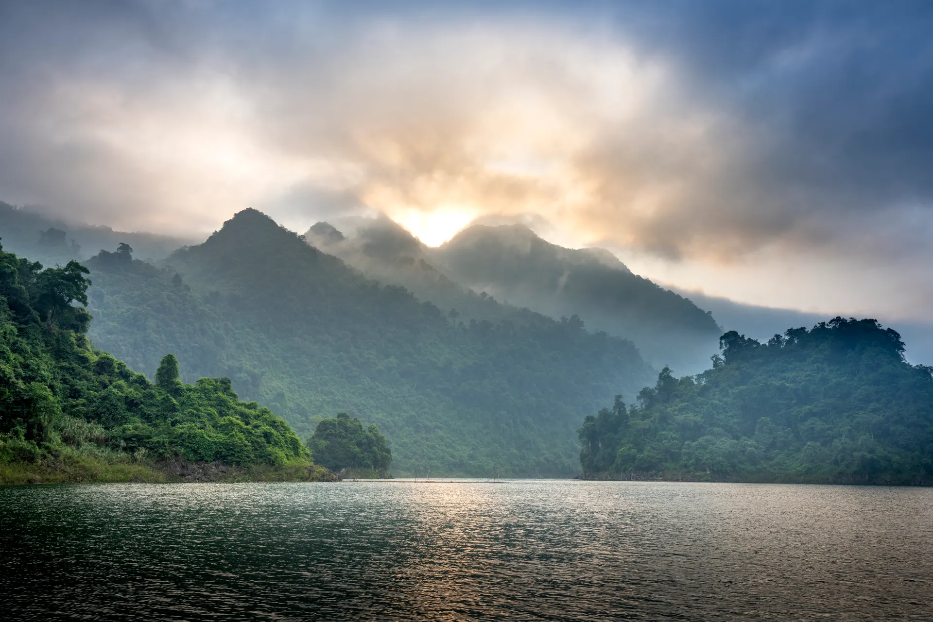 A misty lake ringed by green forested mountains as the sun breaks through at dawn