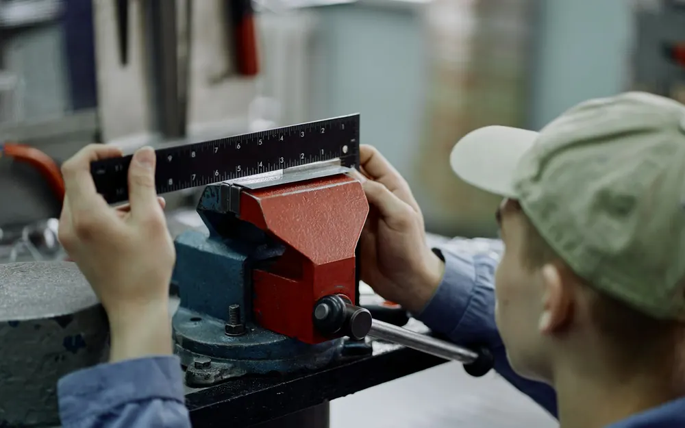 A metal part clamped in a vise being measured against a steel rule