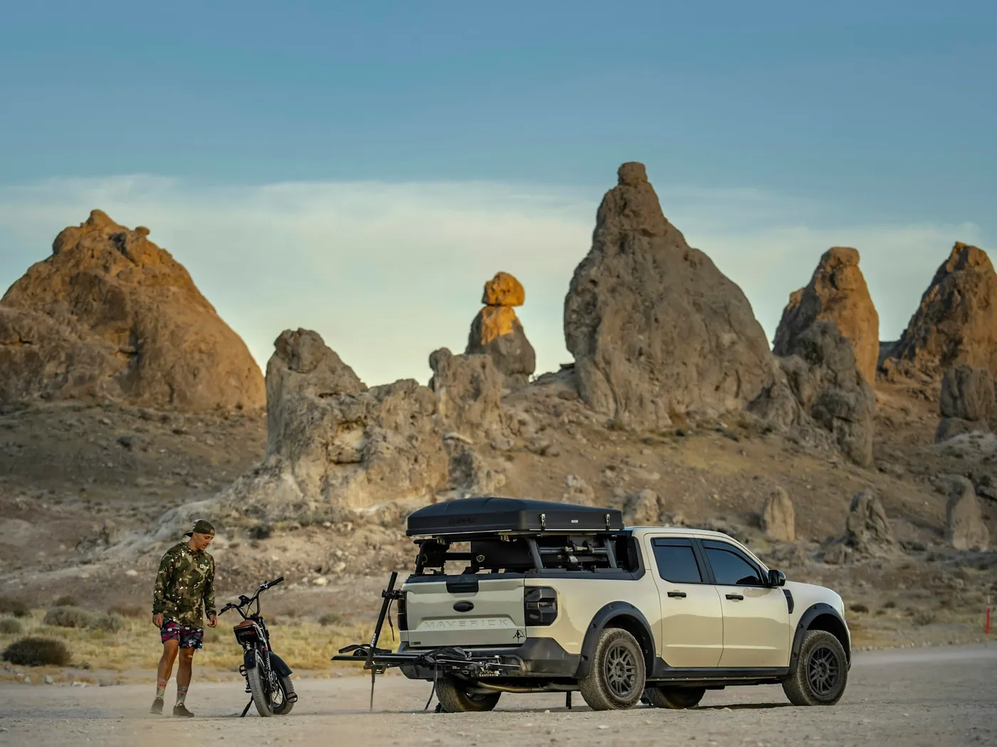 A pick-up with a roof tent and a motorcycle on its rear carrier, parked below desert rock formations